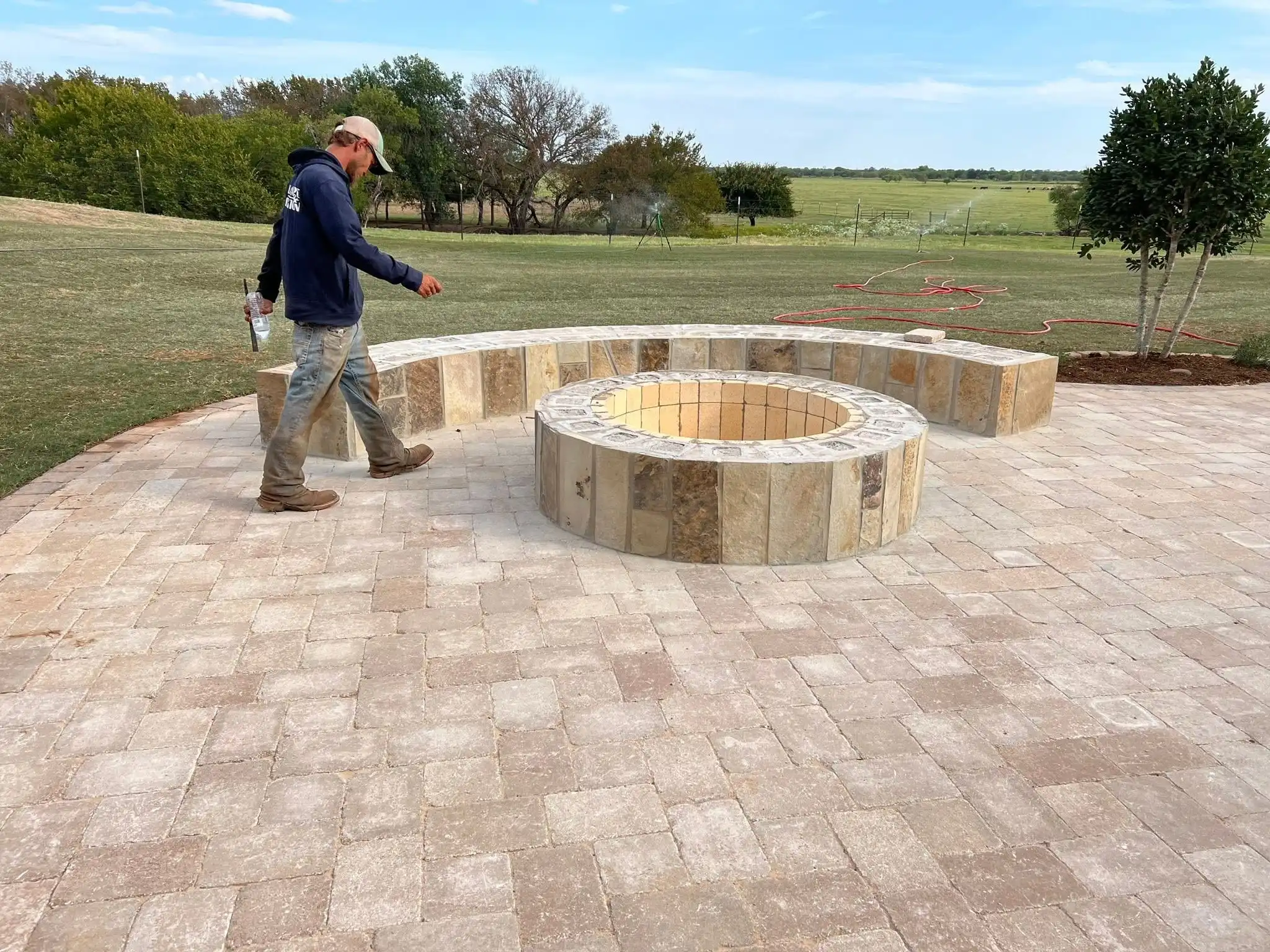 Round paver patio with a stone-faced fire pit and a curved stone seat wall beside a lawn, with a crew member walking across the pavers