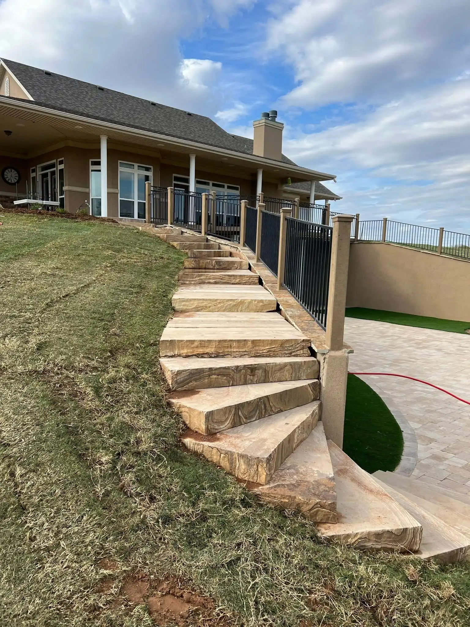 Hillside finished with wide curving stone steps beside a black railing, grass on the slope and artificial turf and pavers at the bottom