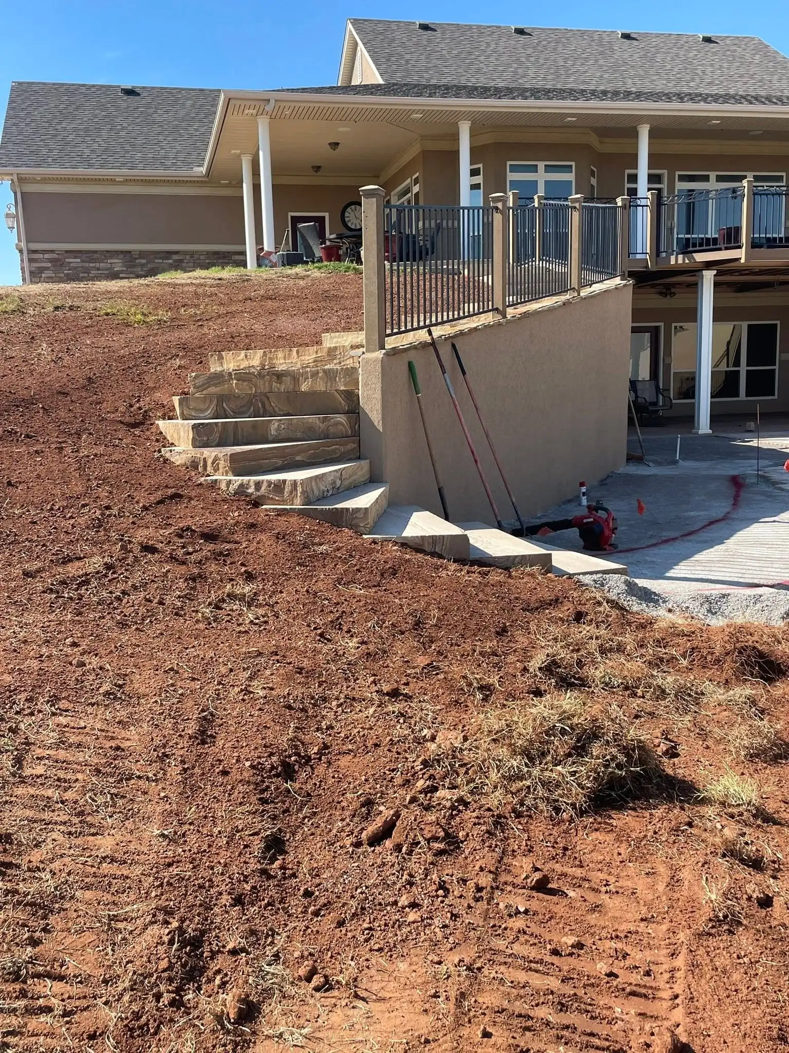 Bare graded red dirt hillside beside new stone steps and a stucco retaining wall, below the back of a home with a deck and railing
