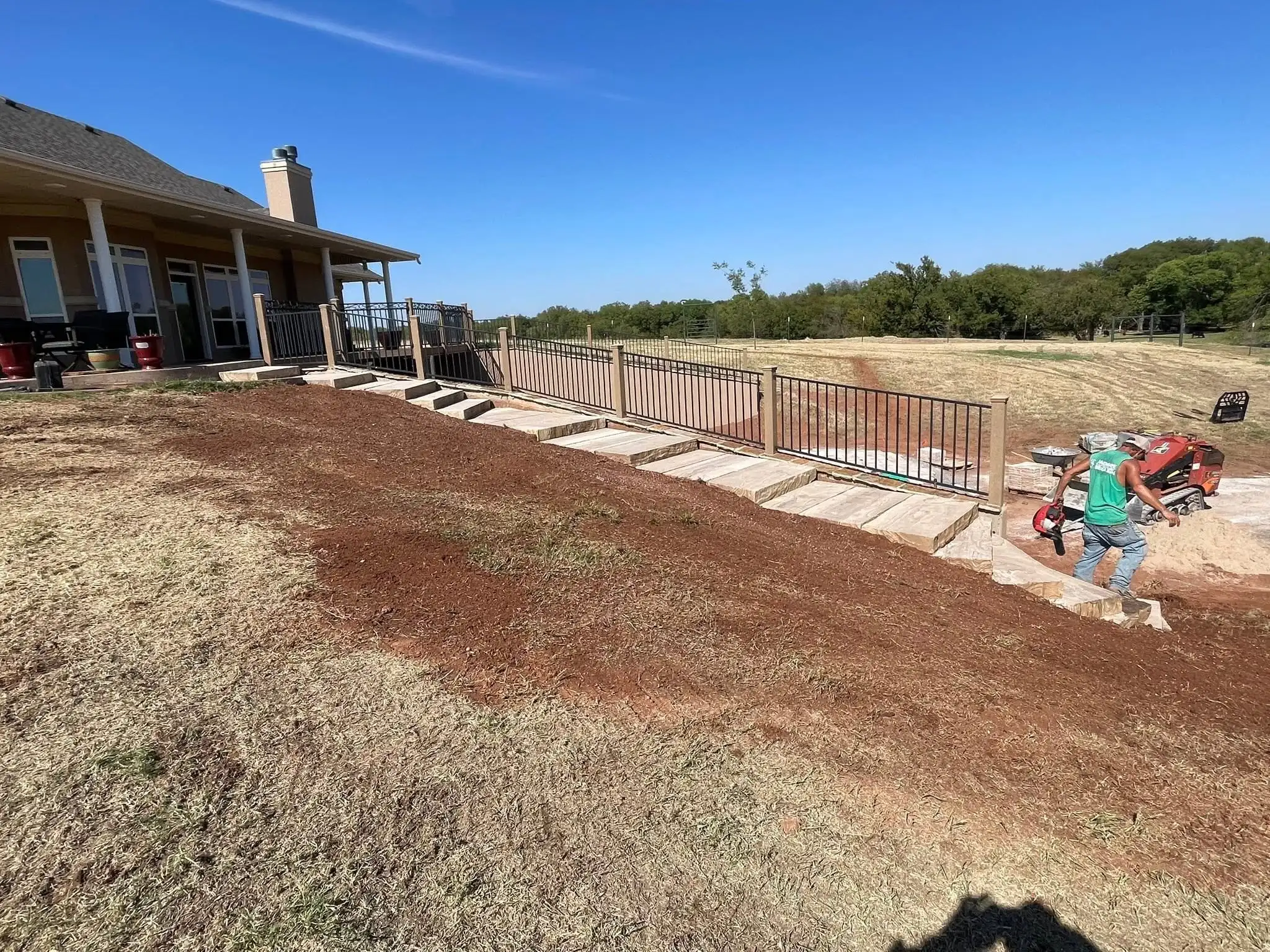 Back slope graded to bare red soil beside a row of stone steps and a black railing, with a crew member carrying a leaf blower and a mini skid steer on the patio base
