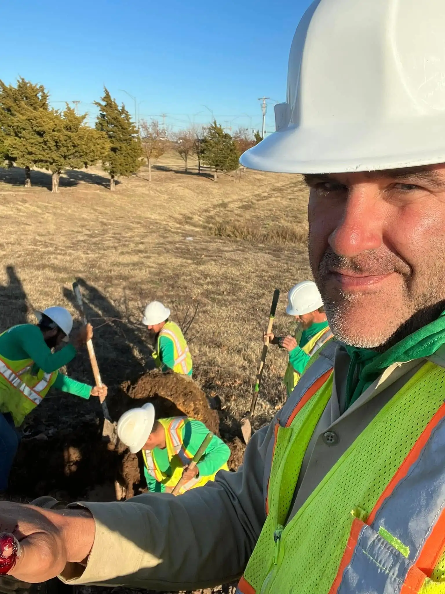 A 4-D leader in a white hard hat and hi-vis vest takes a selfie while four crew members in green shirts, hard hats and vests dig a tree hole behind them