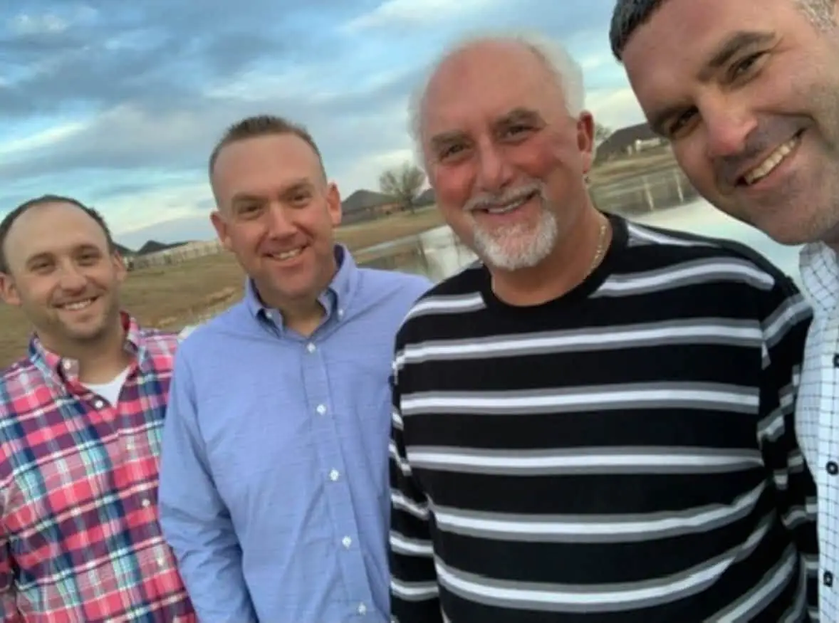 Four men smiling together outdoors in the evening beside a pond