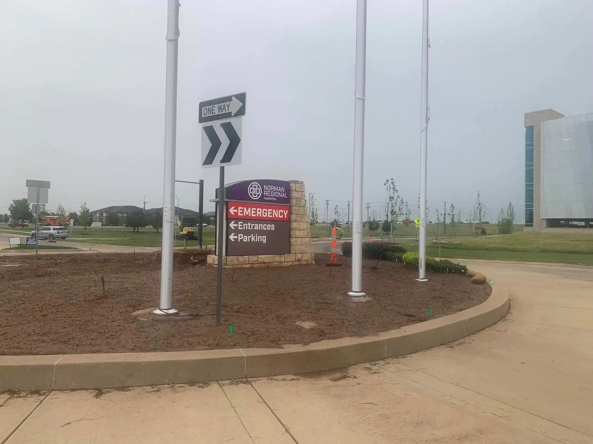 Hospital campus entrance during landscape installation, with fresh soil in a curved sign bed with flagpoles before planting