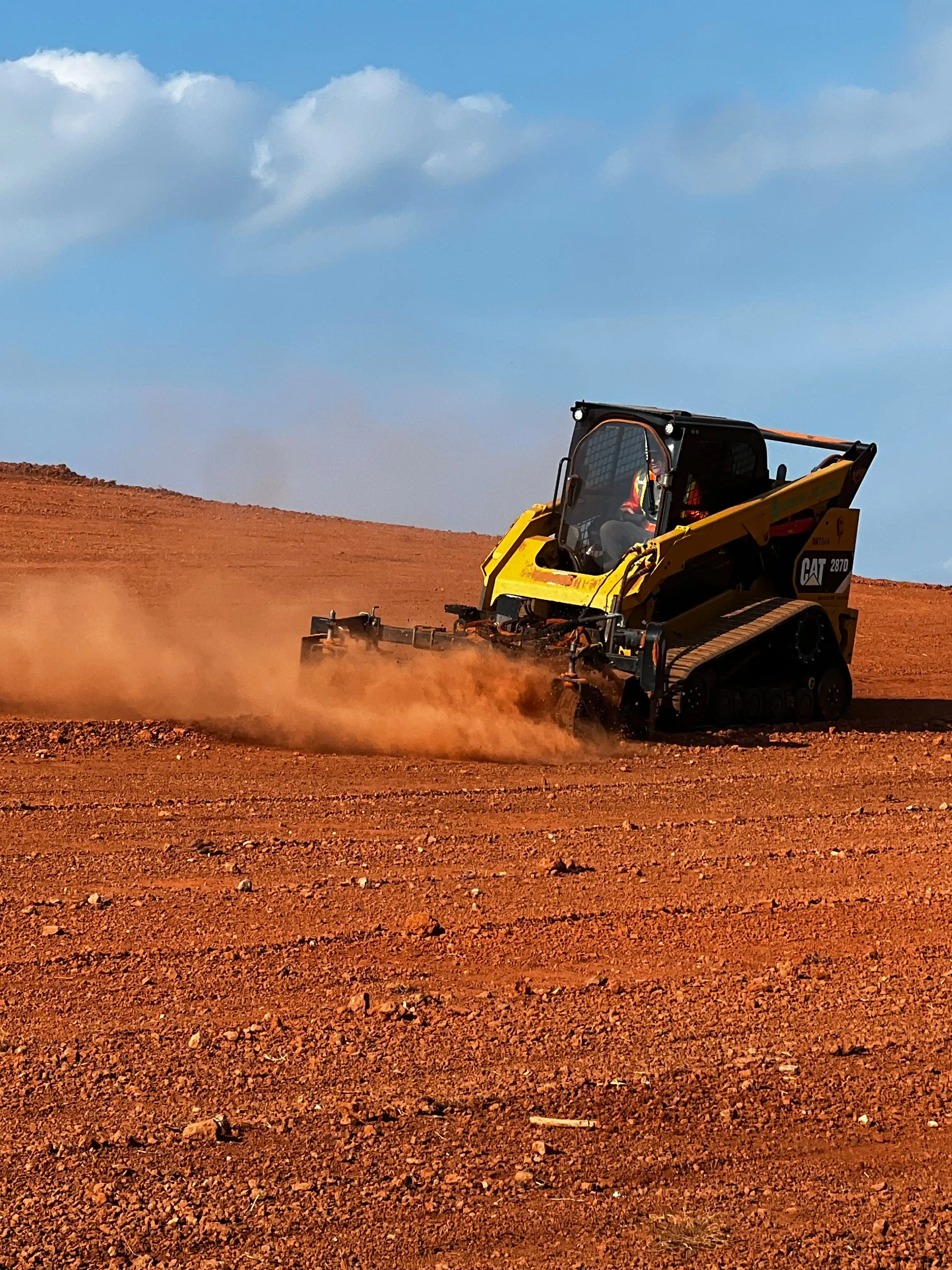 Yellow track loader with a grading attachment smoothing bare red dirt on a large open site, kicking up a cloud of dust under a blue sky