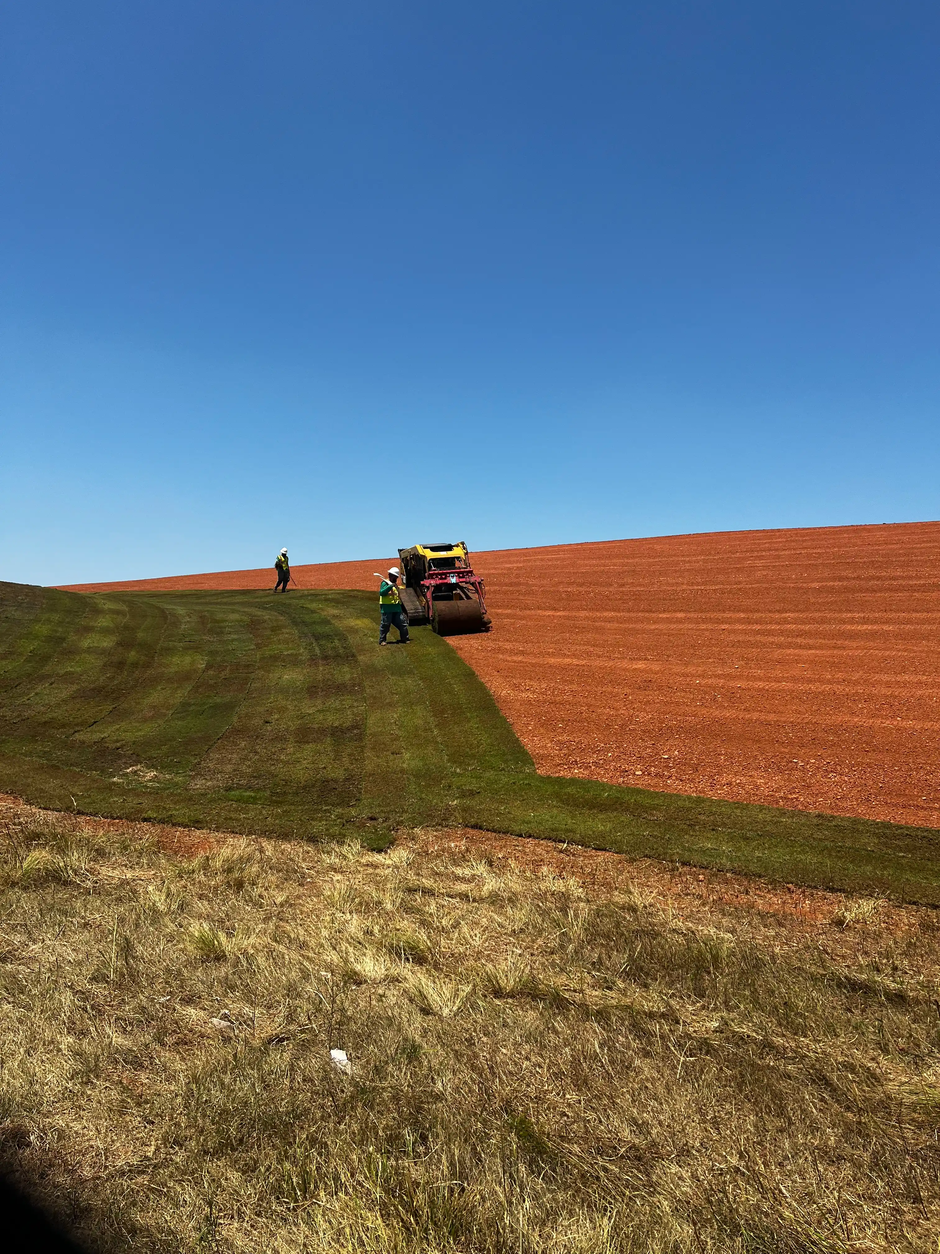 Crew in high-visibility vests laying long strips of green sod with a sod roller across a large red dirt slope under a deep blue sky