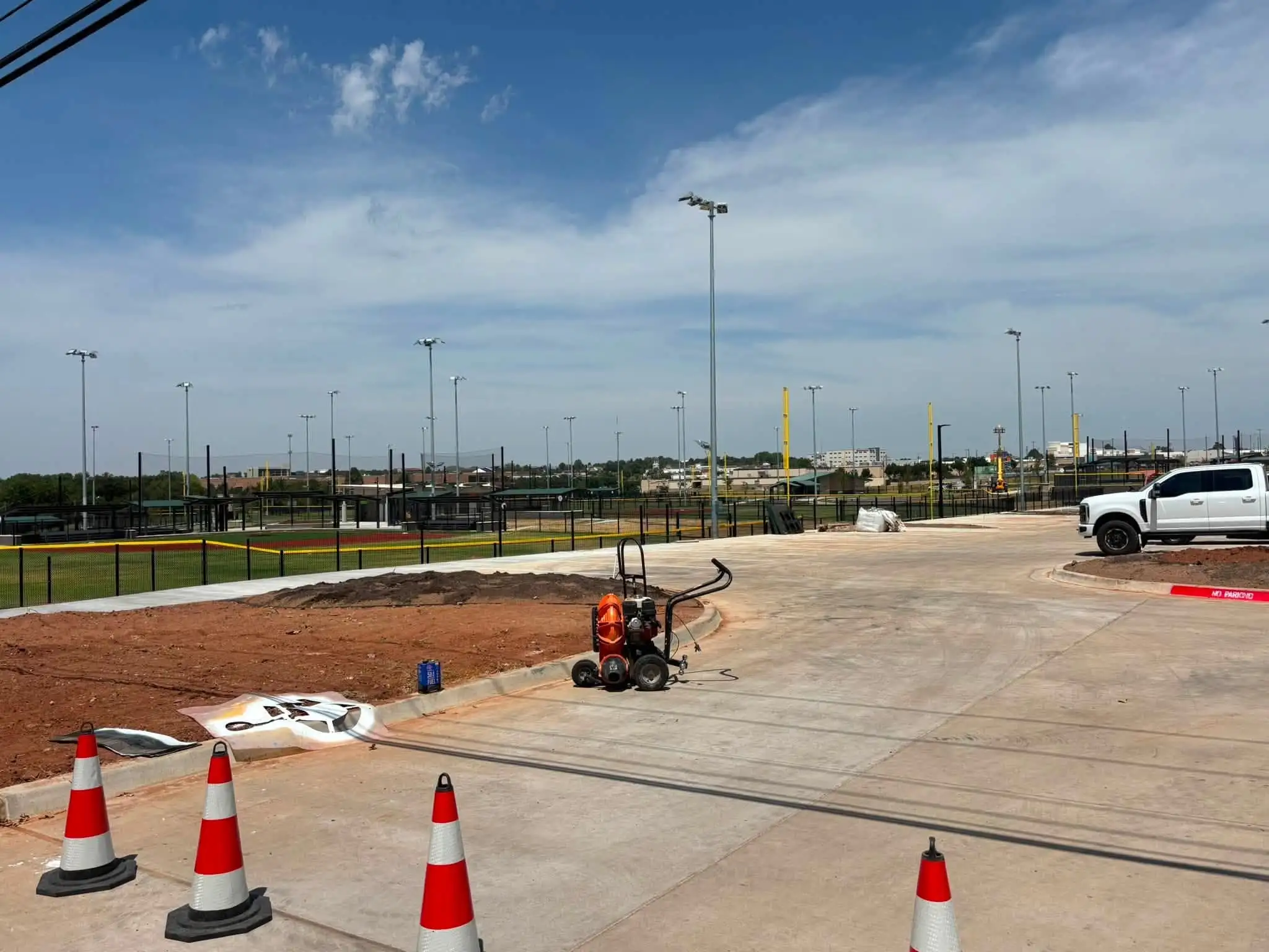 New concrete parking lot with a bare red dirt bed behind the curb, a leaf blower and traffic cones in front, and lit ballfields beyond a black fence