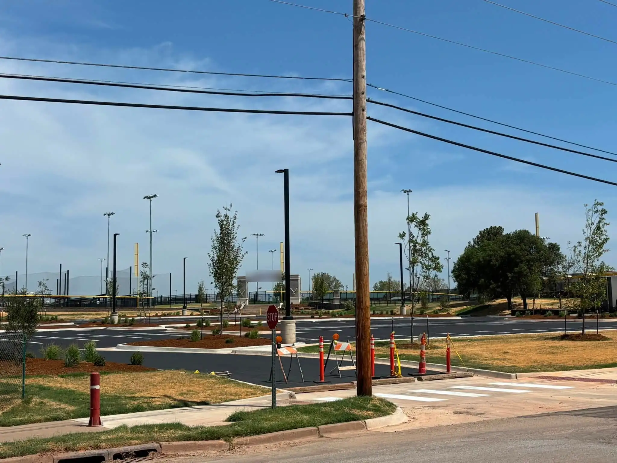 New asphalt parking lot with mulched islands, young trees and ornamental grasses, a striped crosswalk and traffic barricades, with ballfield lights beyond