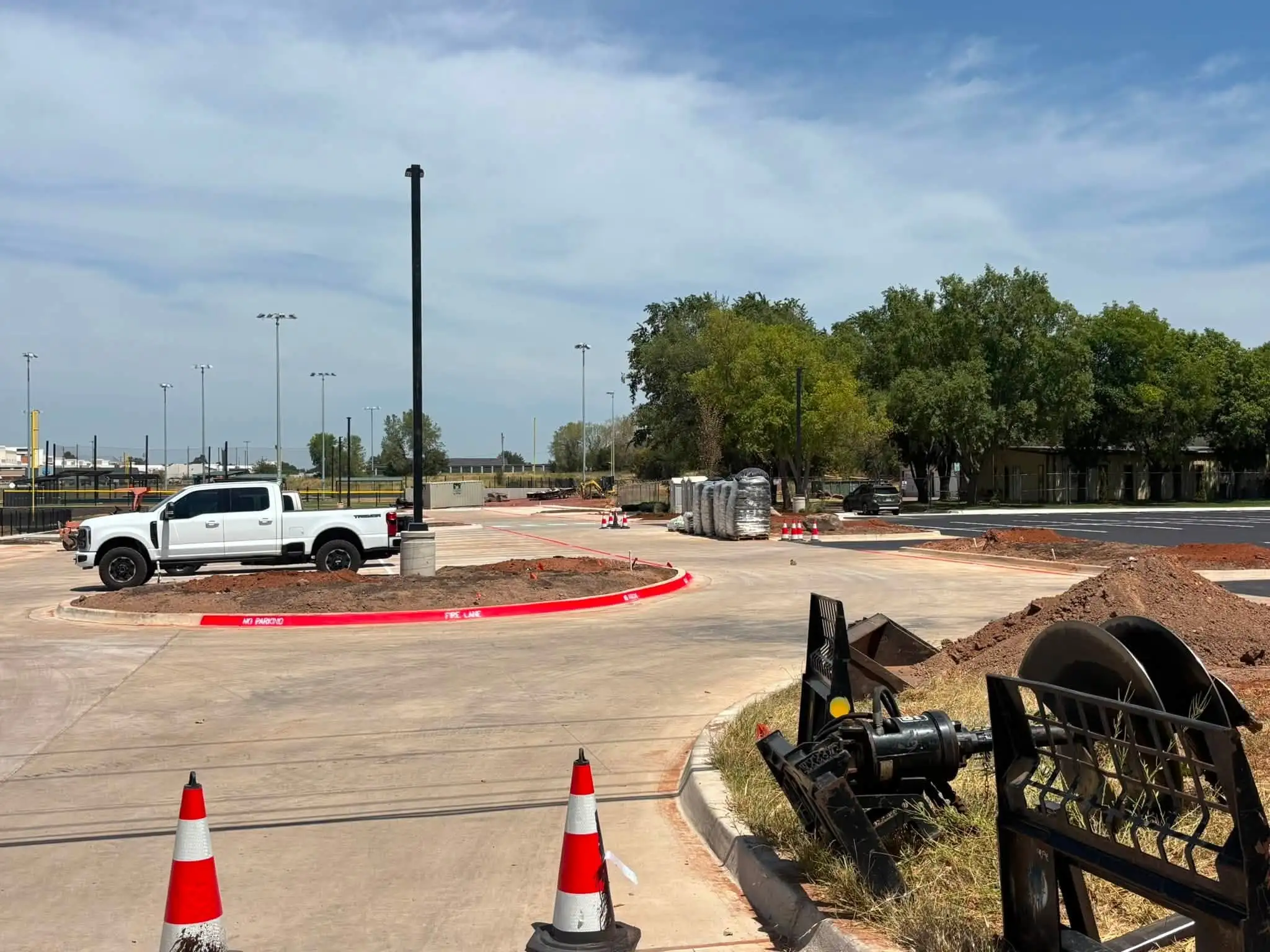 Curving concrete drive around a rounded curb island of bare red soil, with a white pickup, traffic cones, bagged mulch and a skid steer auger attachment