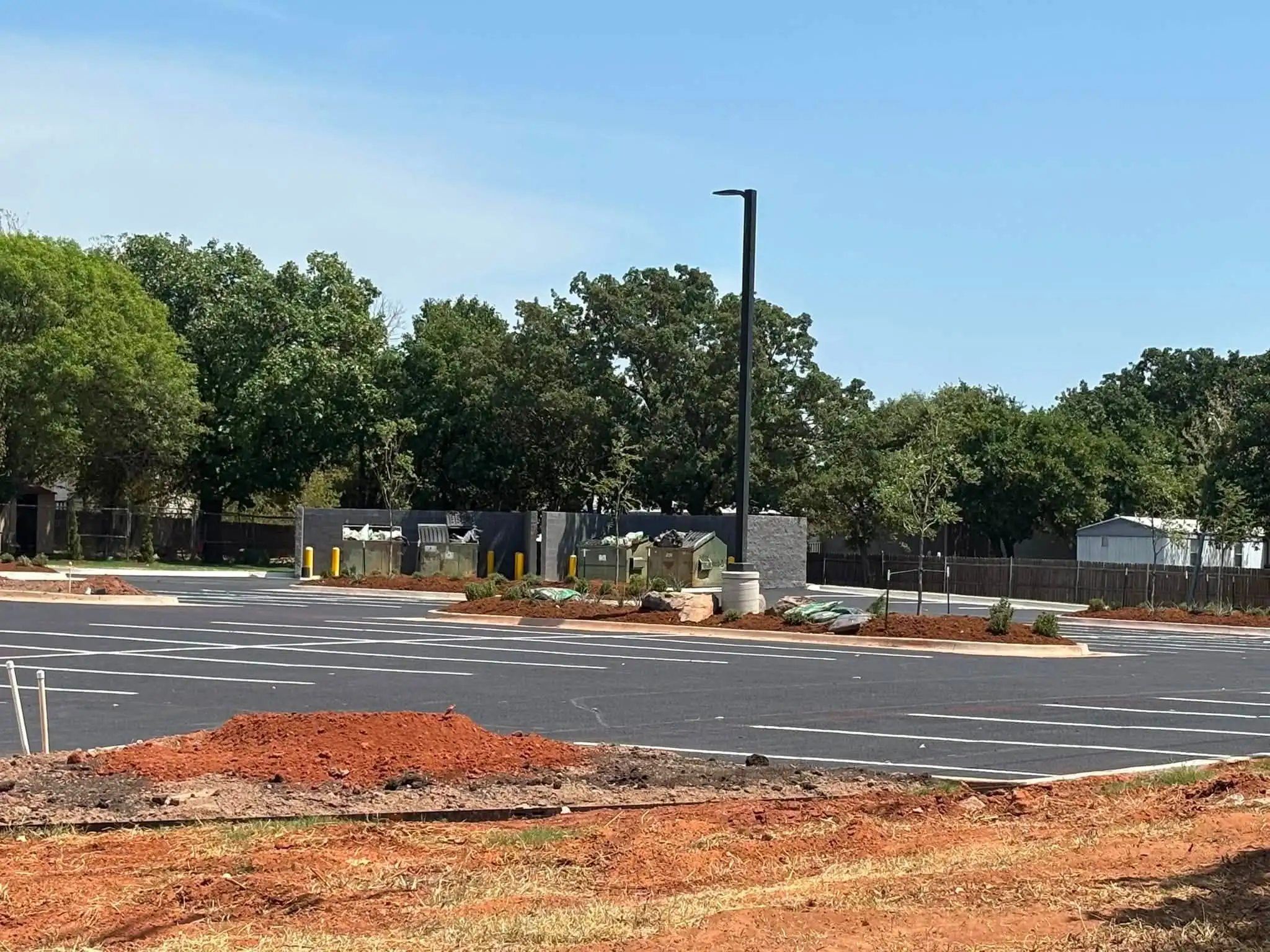 Bare red dirt beds and a mound of soil in the foreground beside a new parking lot, with mulched islands, boulders and young trees already planted beyond