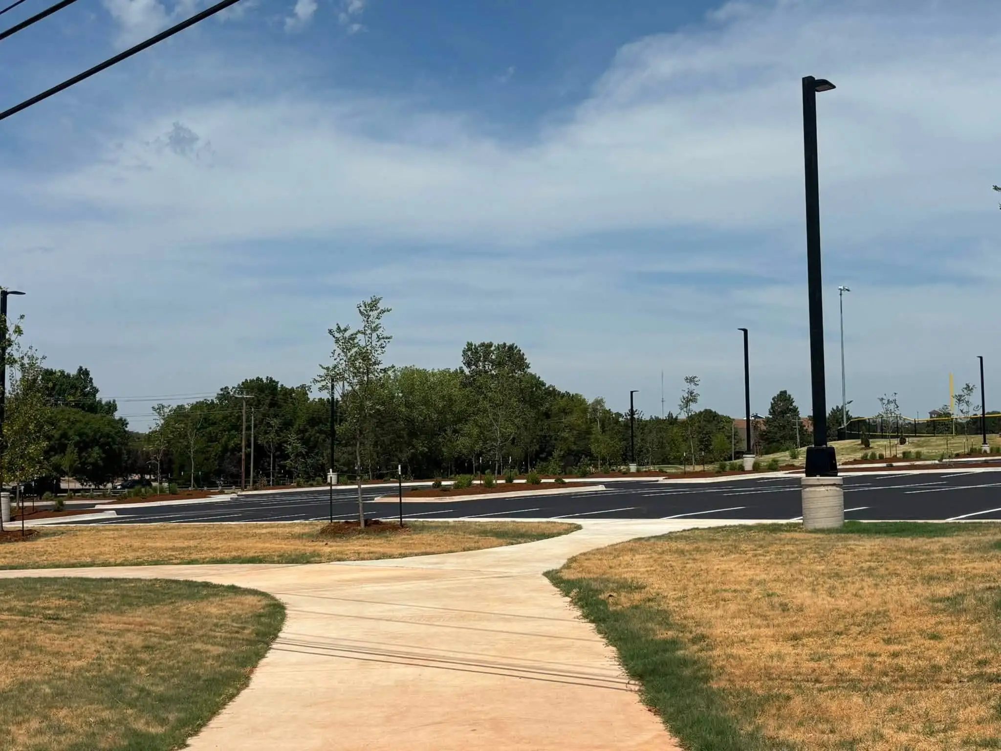 Finished curving concrete sidewalk crossing new turf toward a parking lot lined with mulched beds, shrubs and young trees