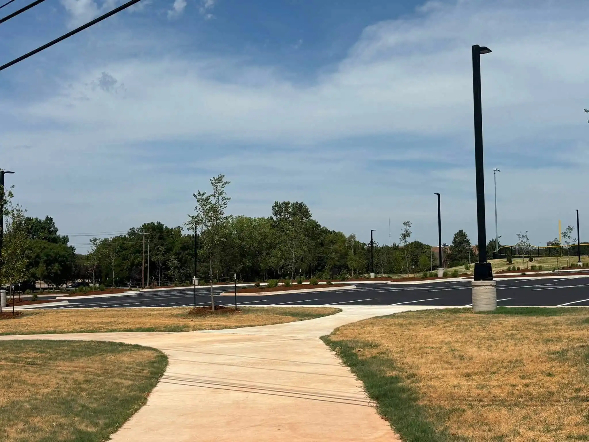 Finished curving concrete sidewalk crossing new turf toward a parking lot lined with mulched beds, shrubs and young trees