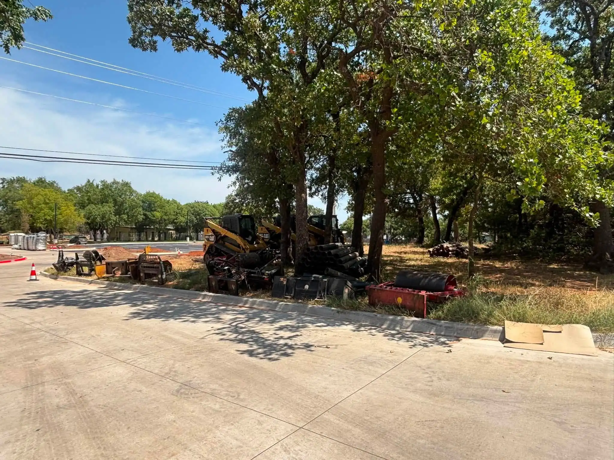 Two yellow compact track loaders, attachments, rolls of black pipe and a red roller parked in the shade of oak trees beside a new concrete road