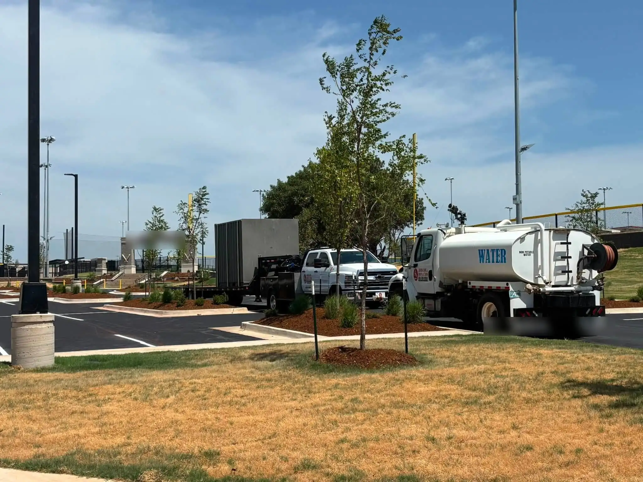 Crew trucks, a trailer and a white water truck parked beside mulched beds with a newly planted tree, fresh turf and a ballfield entrance behind