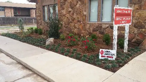 Mulched landscape bed with shrubs along a stone building and a 4-D Lawn and Landscape yard sign