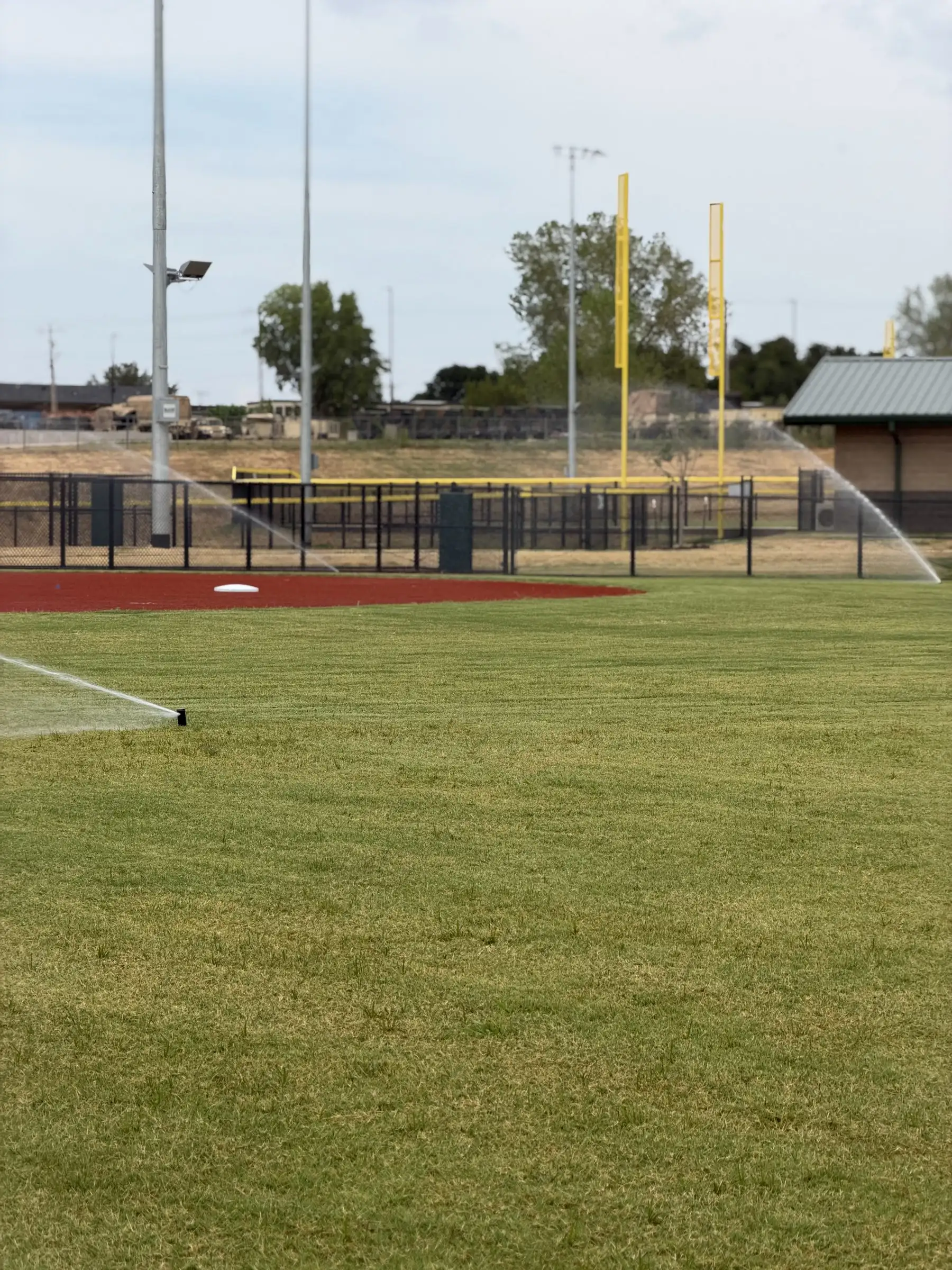 Freshly sodded field beside a ballfield fence with yellow foul poles and covered dugouts beyond