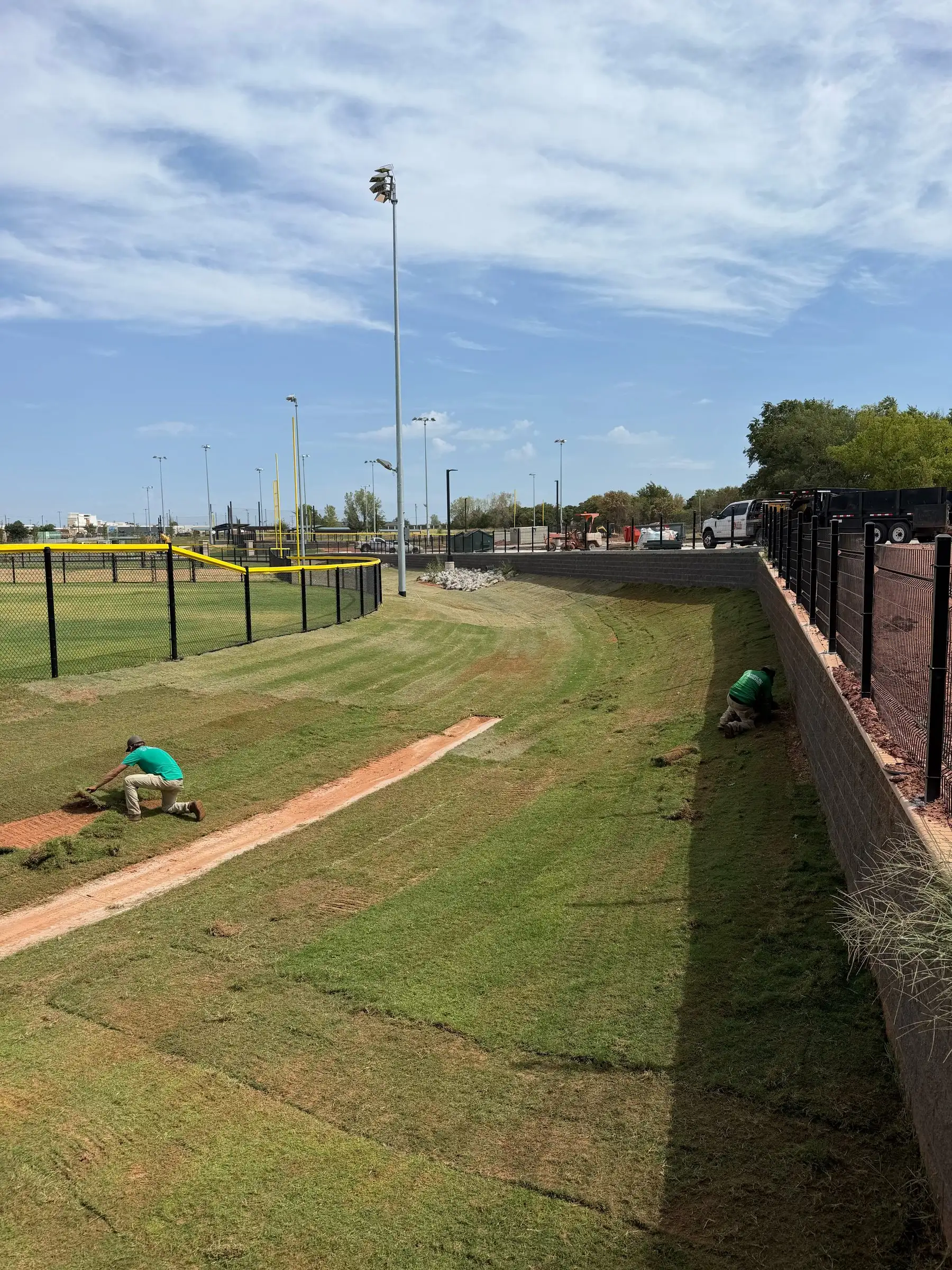 New sod on a slope between a ballfield fence and a block wall, with crew members seen from behind working along a sod seam