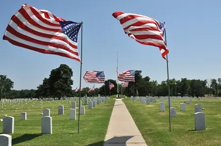 American flags flying along a straight walkway between rows of white headstones on mowed turf