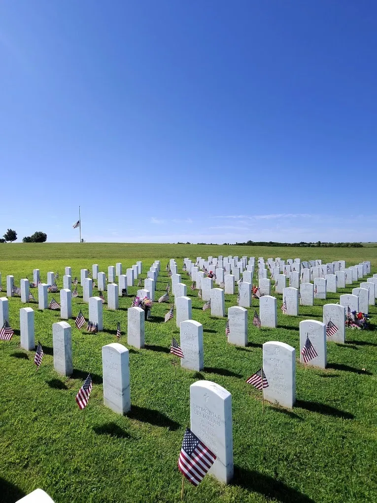 Rows of white headstones with small American flags on green turf at a national cemetery, with a flag at half-staff under a clear blue sky