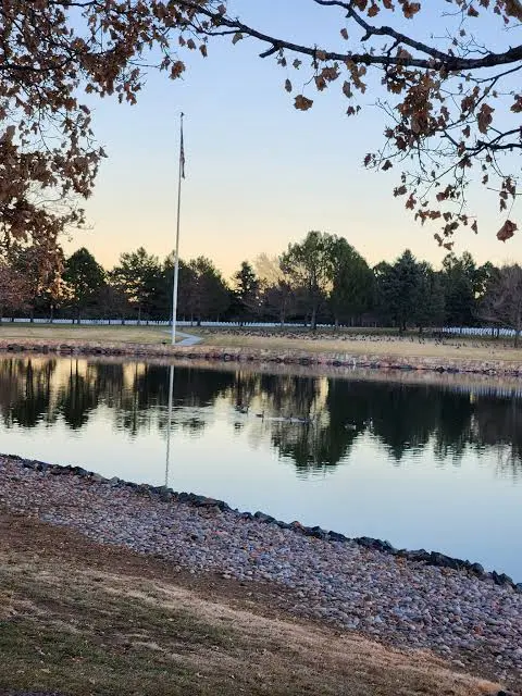 Calm pond at dusk reflecting a flagpole and trees beside the cemetery grounds