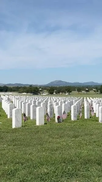 Rows of white headstones with small flags on green turf, with the Wichita Mountains on the horizon