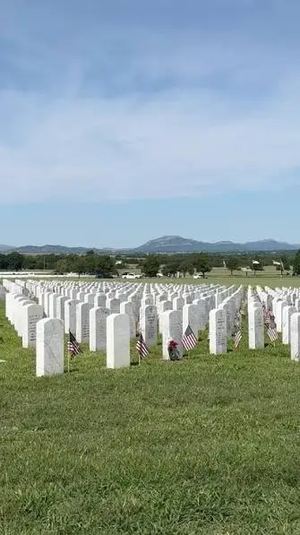 Uniform rows of white headstones with small American flags on mowed green turf at Fort Sill National Cemetery, with the Wichita Mountains on the horizon