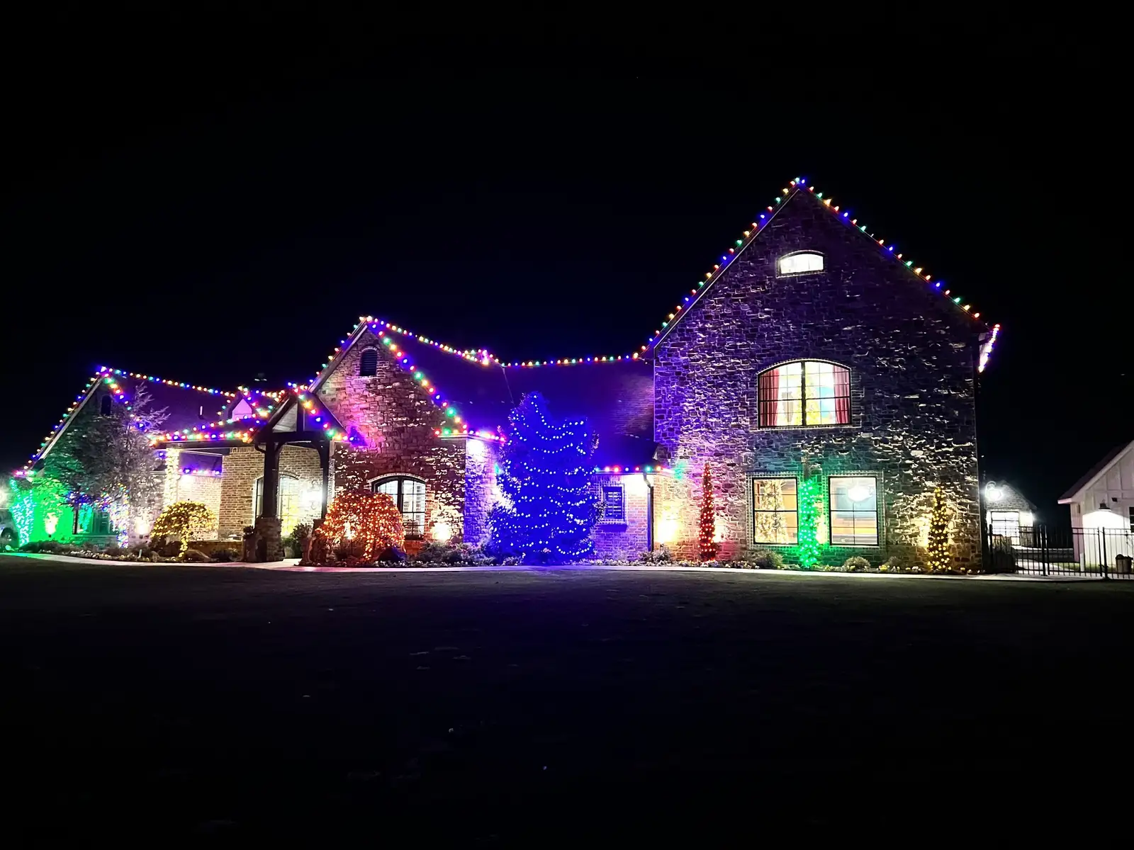 Stone home at night outlined in multicolor holiday lights, with lit trees and shrubs in blue, red, gold and green across the front yard
