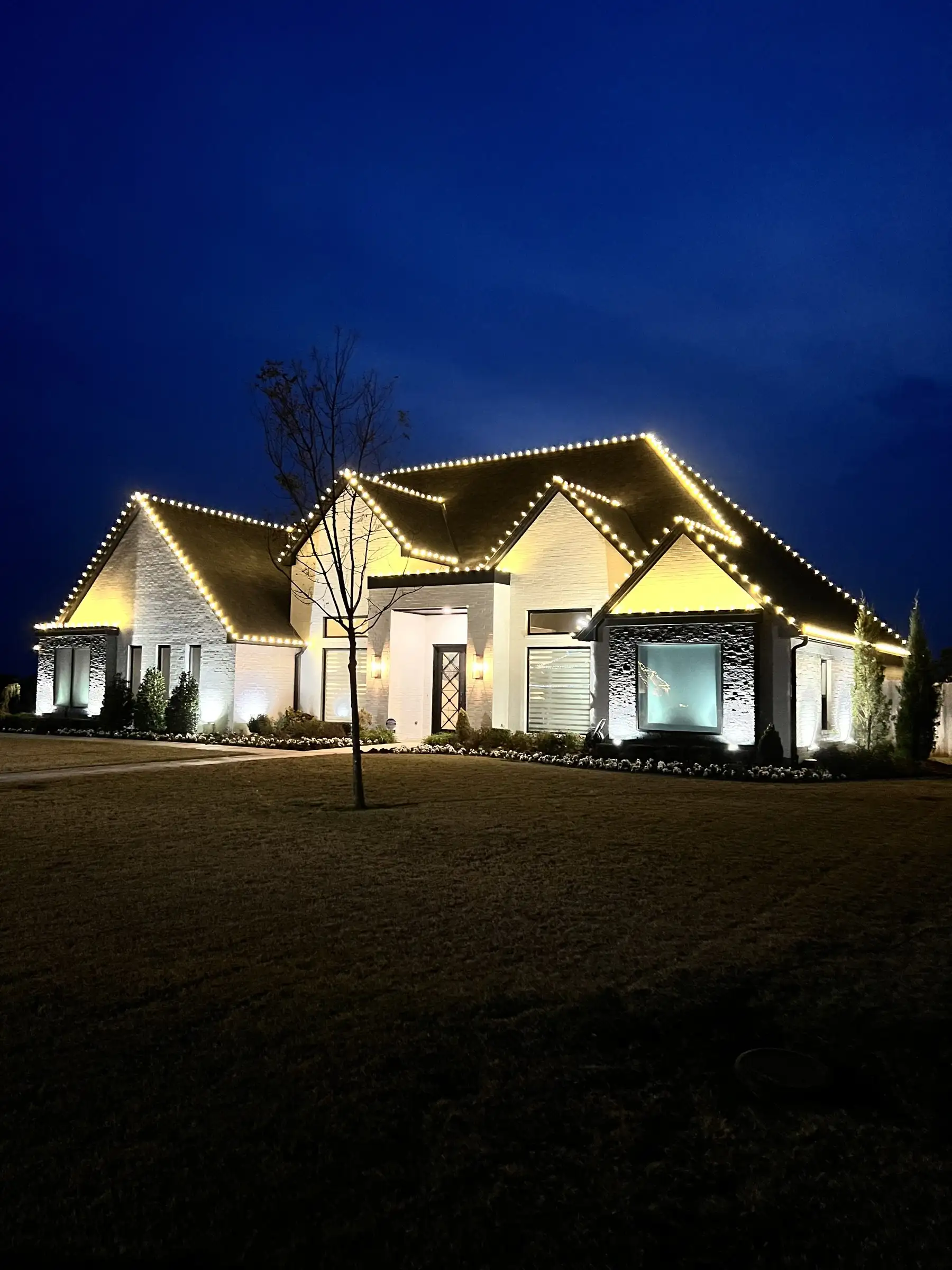 White brick home at dusk with warm white holiday lights outlining every roofline, plus landscape lighting on the walls and beds