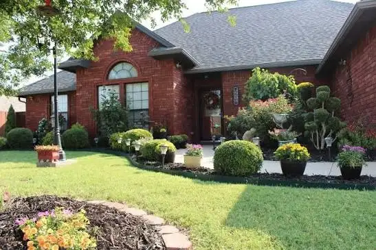 Brick home with a green lawn, fresh mulch beds, round shrubs, topiaries and potted flowers along the walk