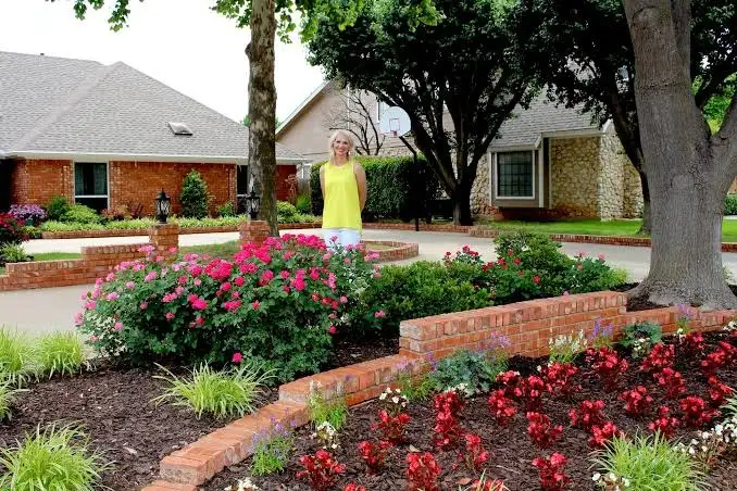 Brick home with rose and begonia beds edged by a brick border, with a woman in a yellow top standing in the yard