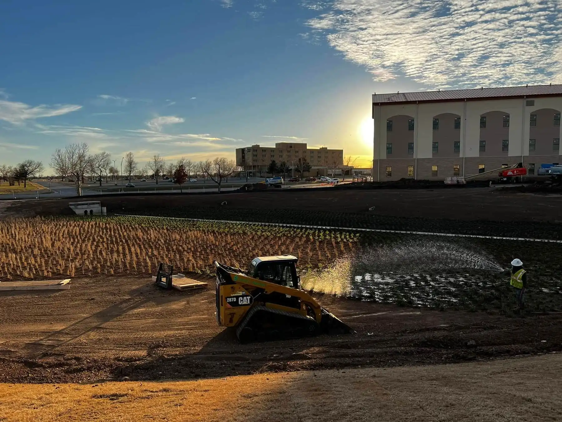 Sunset over a landscape install with a skid steer and fresh plantings being watered by spray